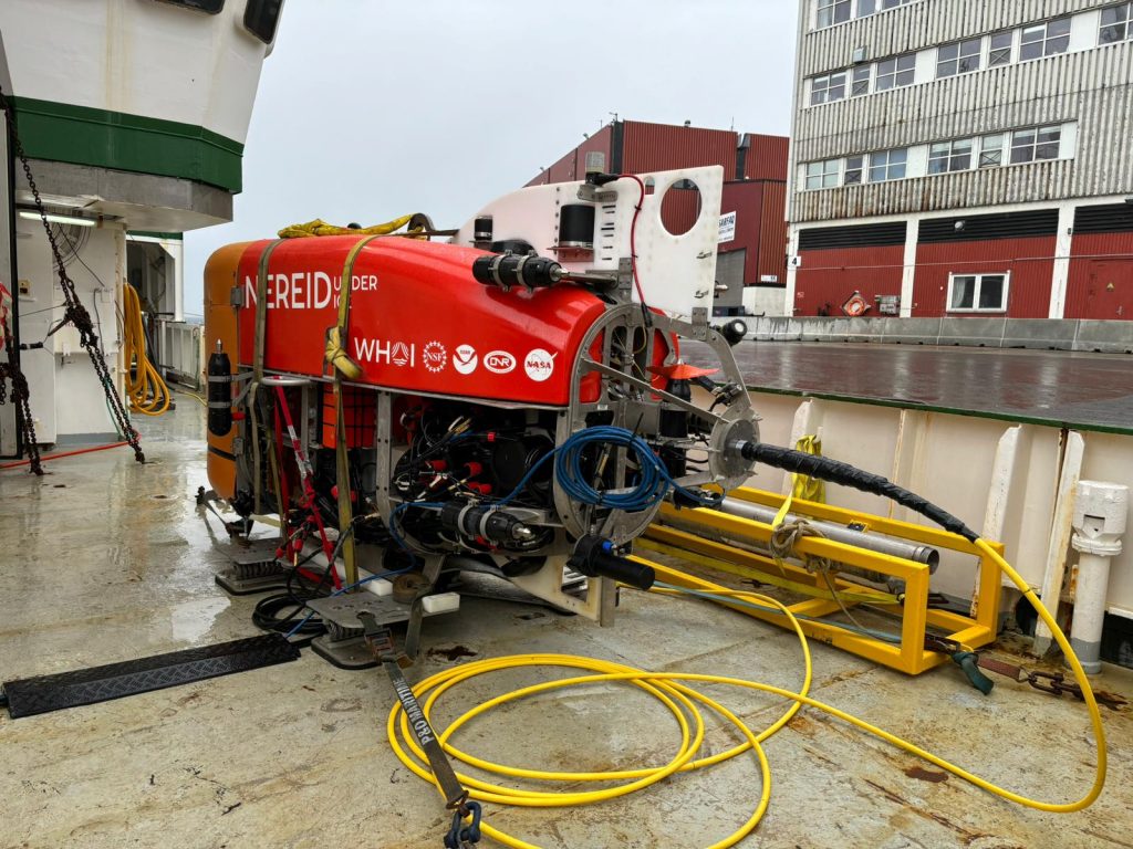 The car sized red submersible sits on the ship's deck while docked.