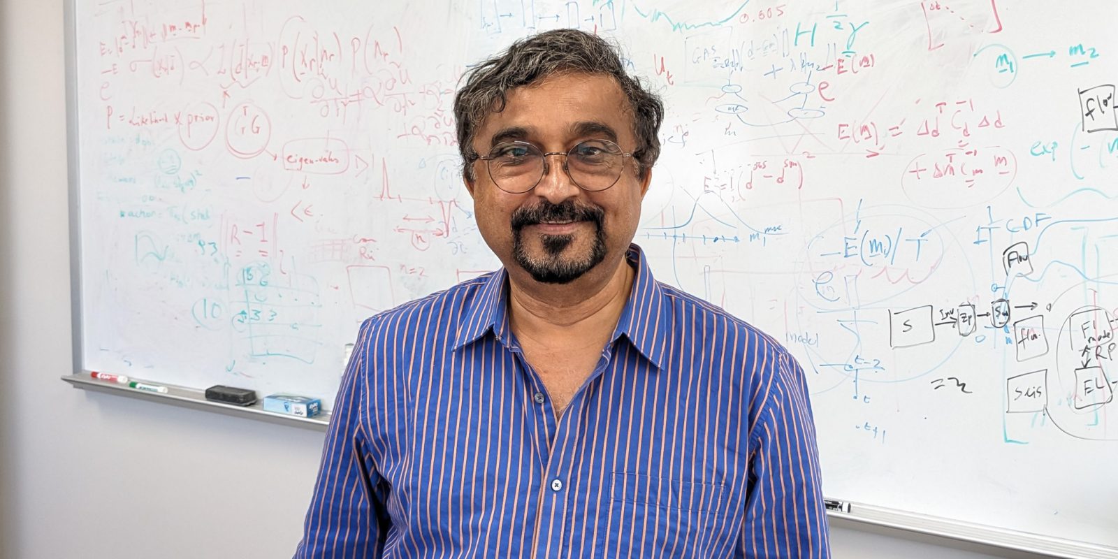 Photo of Mrinal in his office in front of a strategically placed whiteboard with equations.