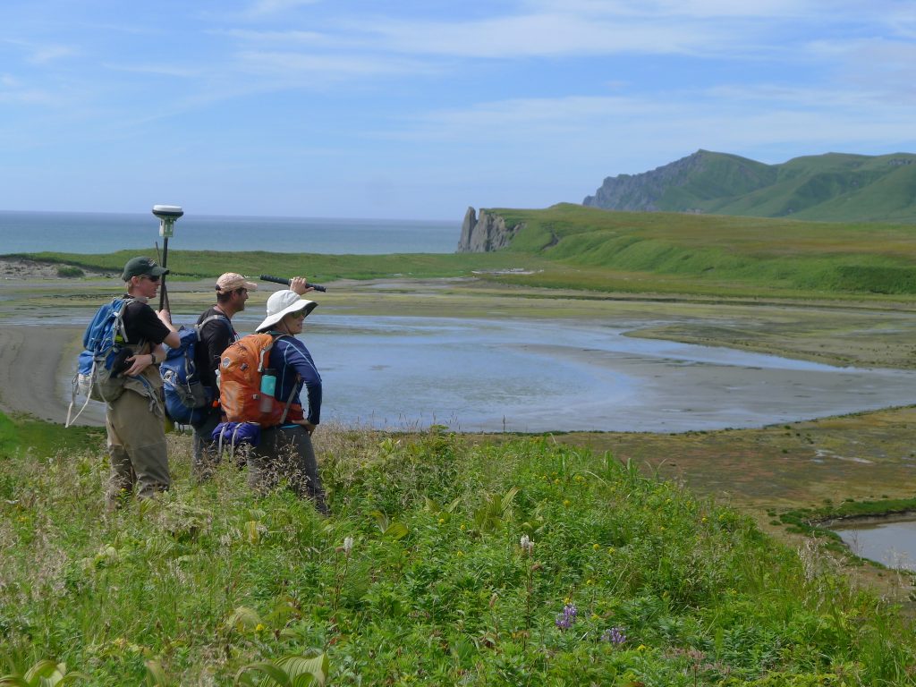 A photo of Tina and two other researchers, standing on a green field looking out to a body of water.