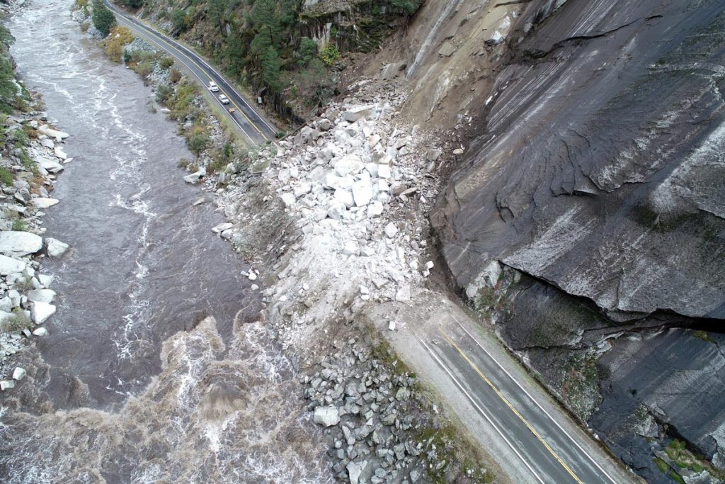 Aerial view of a landslide blocking a road next to a river, with debris covering both lanes and encroaching on the riverbank.