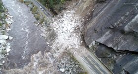 Aerial view of a landslide blocking a road next to a river, with debris covering both lanes and encroaching on the riverbank.