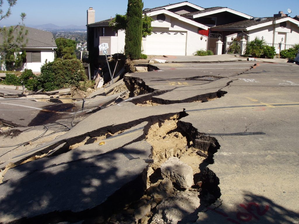 A large crack runs the entire width of a residential road with the road on the left side of the image substantially lower than the other.