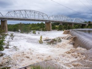 Photo of swollen river overflowing a dam as it runs under a truss bridge. The river is fast flowing and brown.