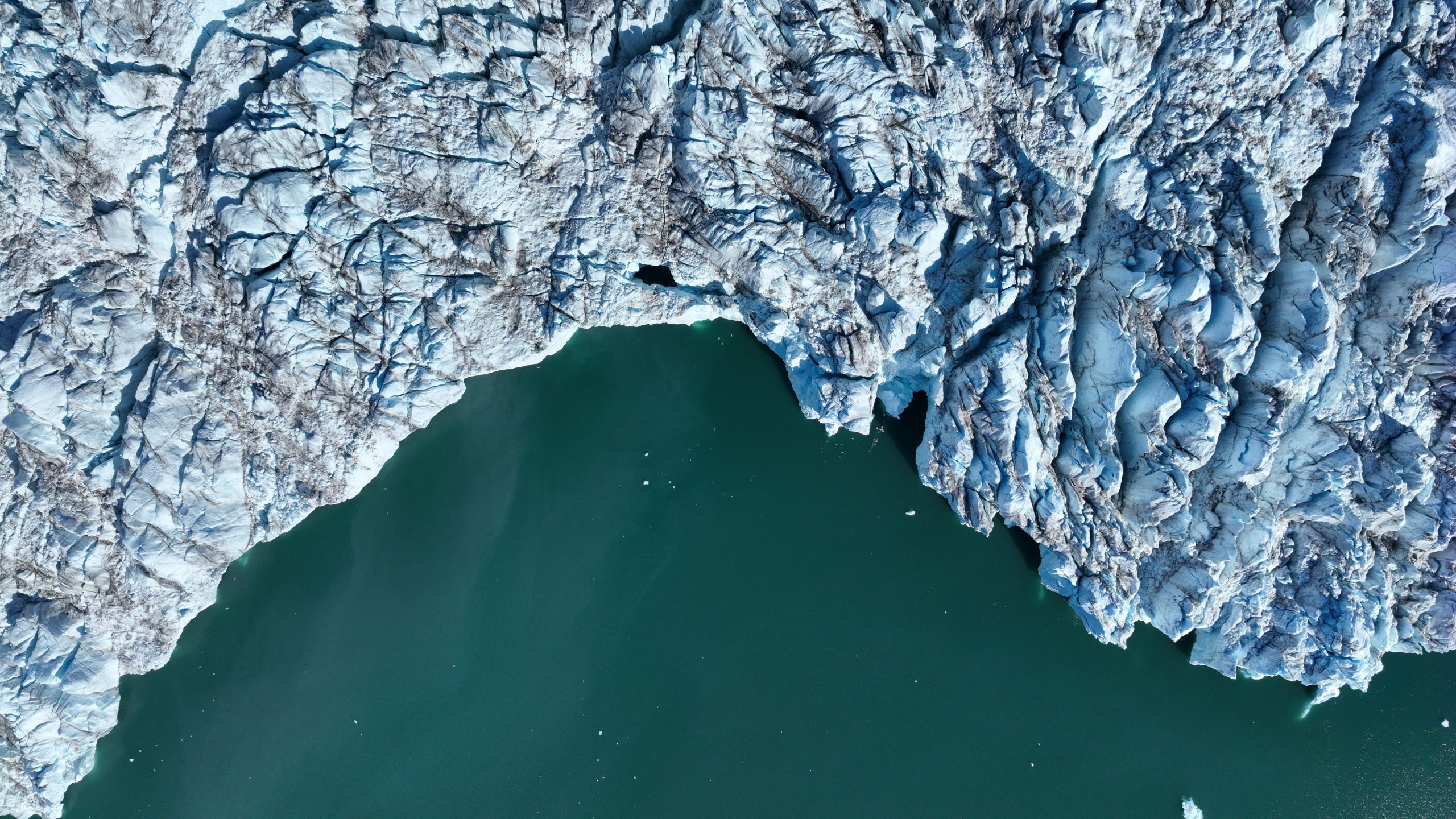 Top down view of the edge of a glacier. The sea is a dark green, the ice dirty blue and textured like elephant skin.