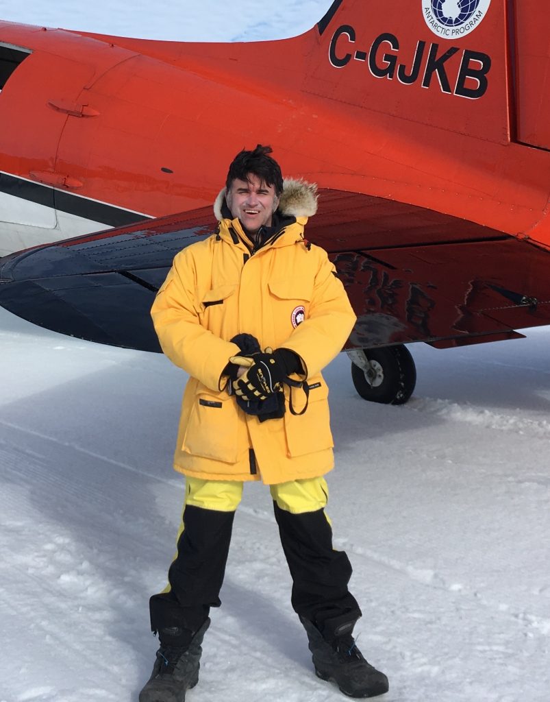 Photo of Duncan in yellow polar gear posing for a photo in front of a red DC-3 parked on the ice.