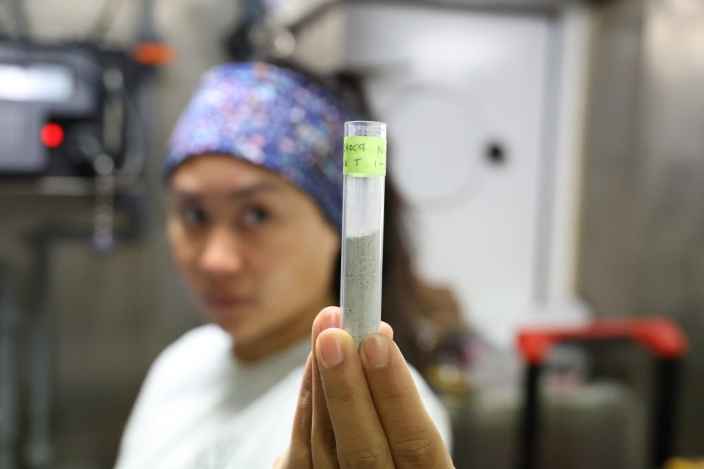 Close up of a small test tube half filled with grey powder. It's held by Mikayla.