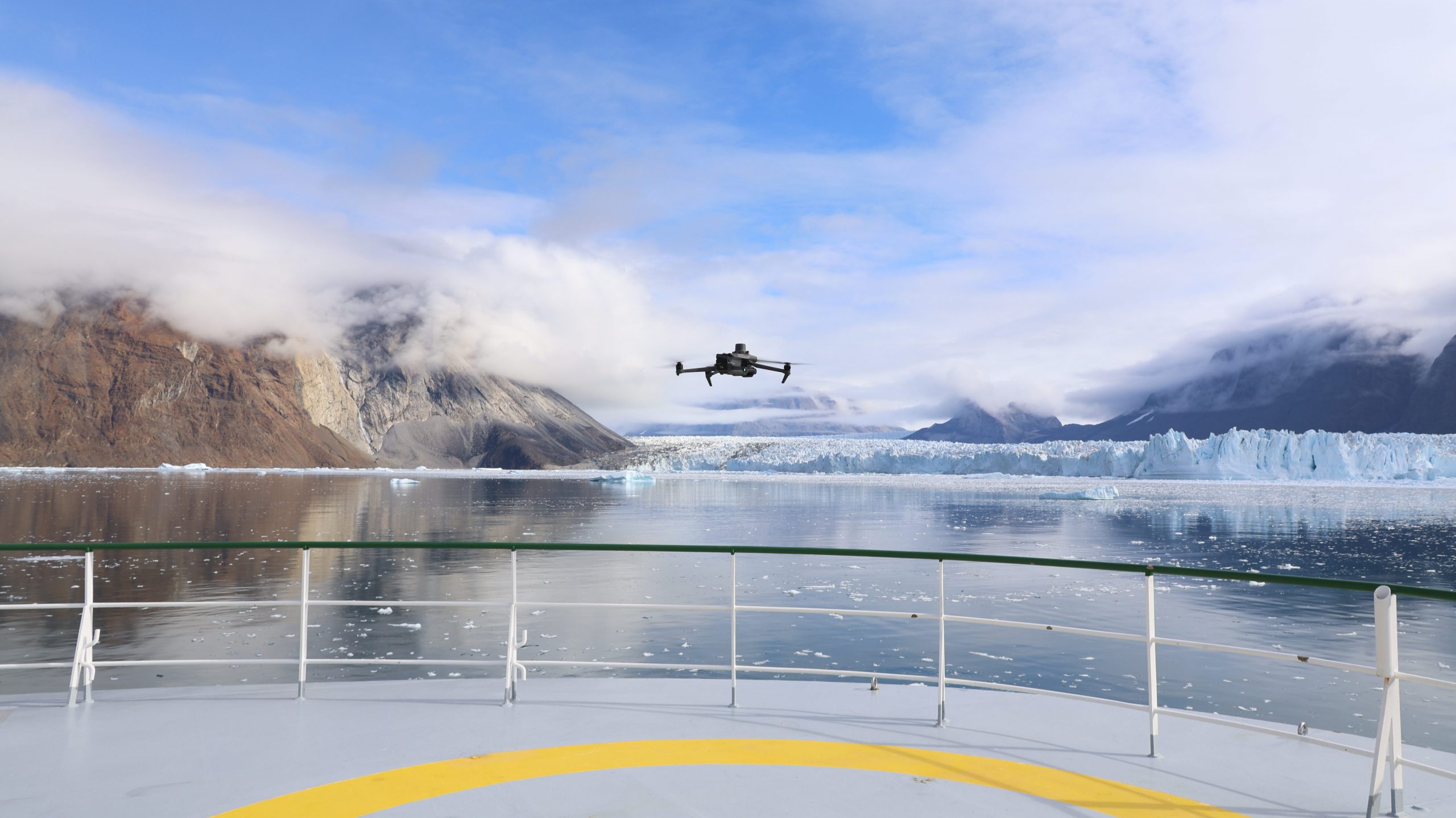 Photo of the drone hovering over the ship's bow with the glacier behind it.