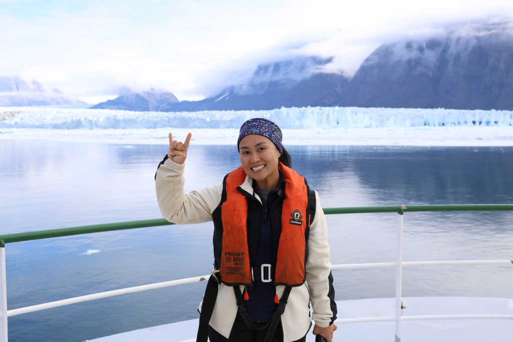 Photo of Mikayla flashing a hook 'em on the bow of the ship with the glacier behind her. She is wearing a life vest.