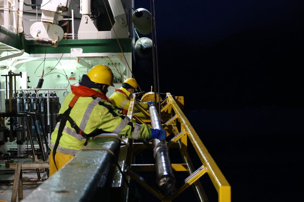 Deck crew hauling a metal cylinder from the side of the vessel at night.