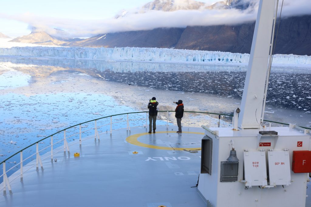 Photo looking down on the ships bow, where two people are looking out over the sea. The glacier is in the distance.