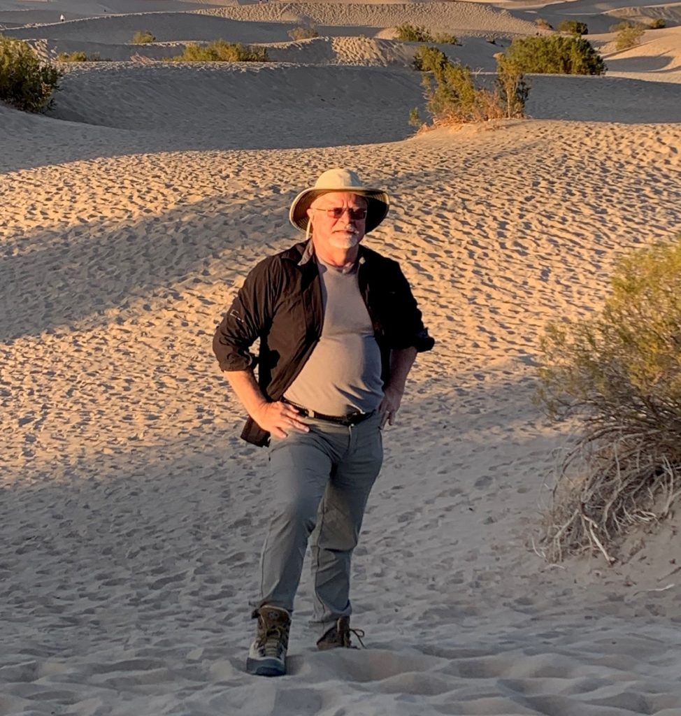 Photo of Tim standing in a sandy desert, wearing a hat.