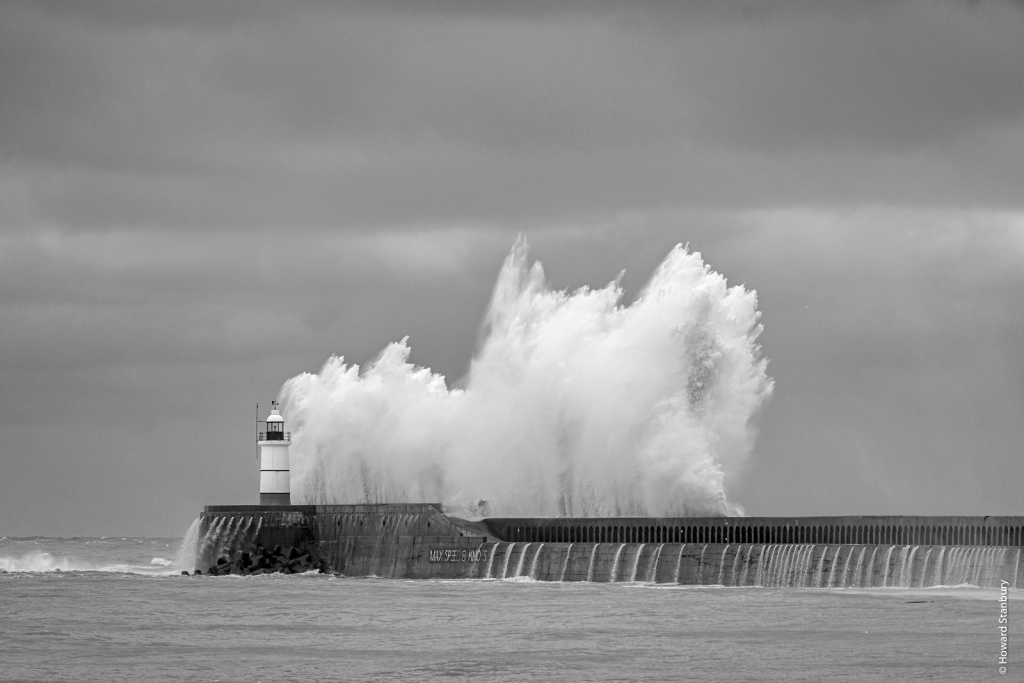 Black and white photo of a large storm wave crashing against a lighthouse and wave break beneath stormy skies.