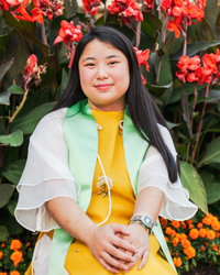 Photo of Mandala wearing a yellow dress and a mint green graduation sash. She is standing in front of a garden with red flowers.