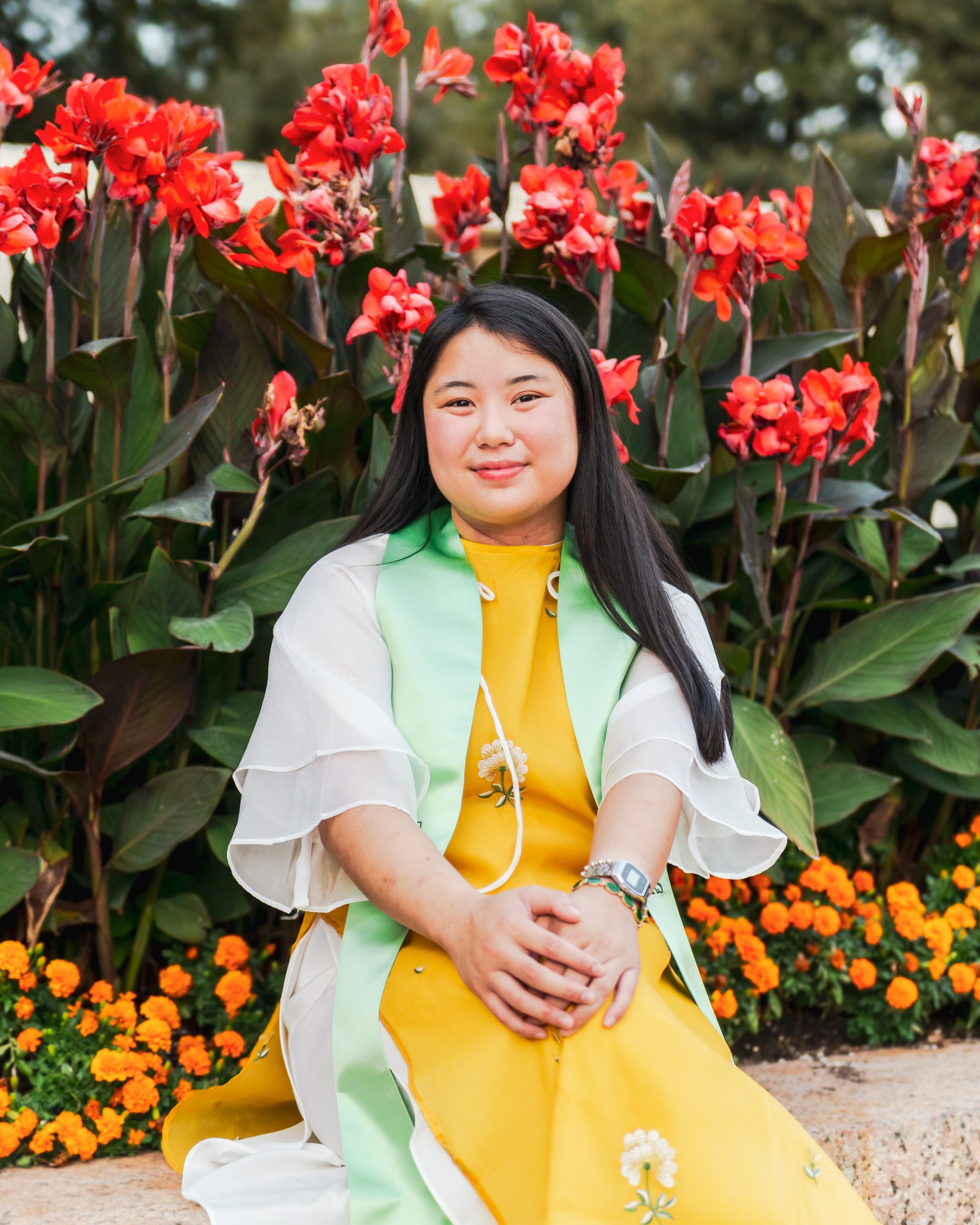 Photo of Mandala wearing a yellow dress and a mint green graduation sash. She is standing in front of a garden with red flowers.