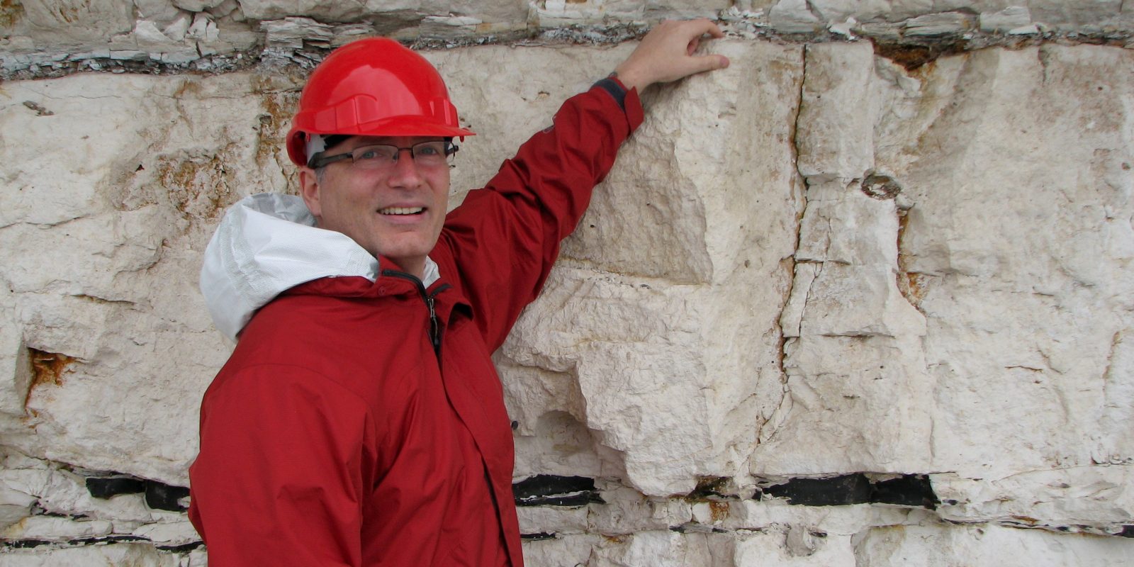 Photo of Richard in a red hard hat and jacket pointing at a dark line on a walk of chalk or limestone.