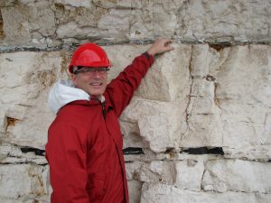 Photo of Richard in a red hard hat and jacket pointing at a dark line on a walk of chalk or limestone.