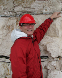 Photo of Richard in a red hard hat and jacket pointing at a dark line on a walk of chalk or limestone.