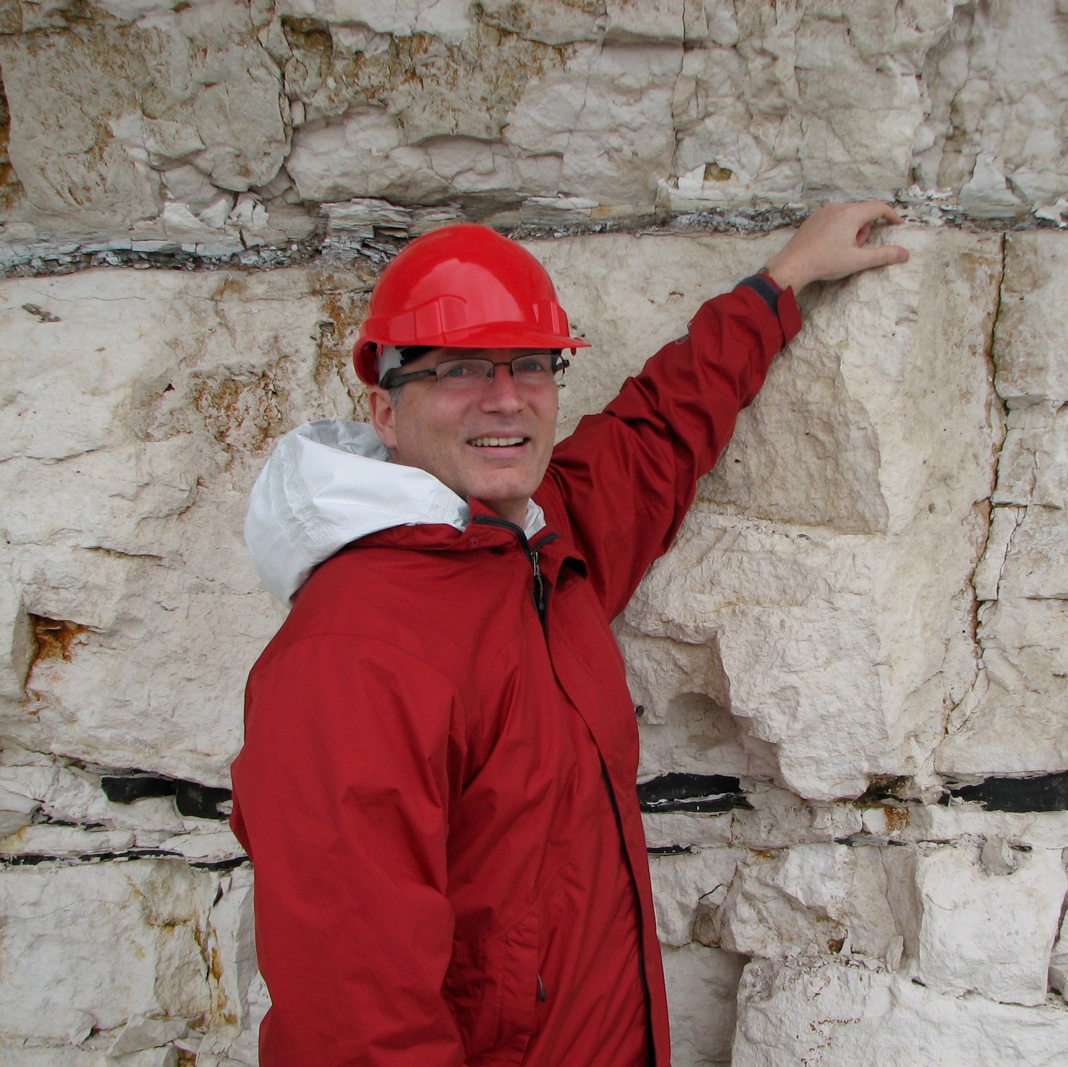 Photo of Richard in a red hard hat and jacket pointing at a dark line on a walk of chalk or limestone.