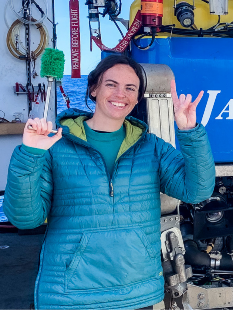 Photo of Kaitlin. She is wearing a blue jacket, and is standing in front of a remotely operated vehicle (ROV) system.